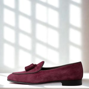 May include: A burgundy suede loafer with a tassel detail. The shoe has a rounded toe and a black sole. The shoe is approximately 11 inches long.