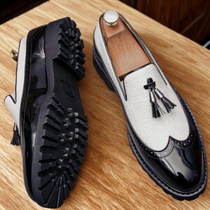 May include: A pair of two-tone loafers with a black and white design. The shoes feature a textured white upper, black patent leather accents, and decorative tassels. The thick black soles have a rugged tread.