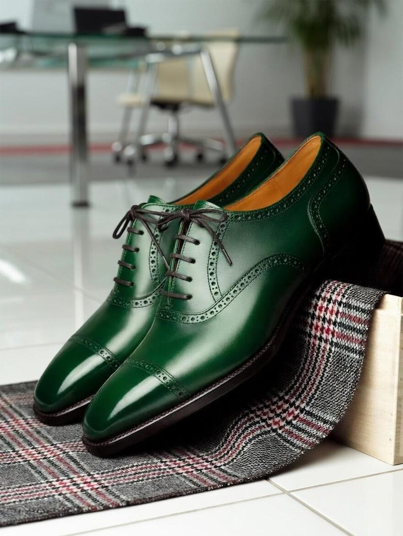 May include: A pair of polished, emerald green leather dress shoes with brown laces. The shoes feature a classic cap-toe design and decorative perforations. They rest on a patterned fabric, suggesting a focus on formal footwear.