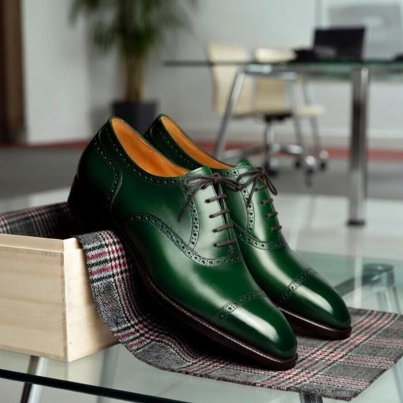 May include: A pair of polished, emerald green leather dress shoes with brown laces. The shoes feature a classic wingtip design with decorative perforations. They are displayed on a plaid fabric and a wooden box, suggesting a high-end product.