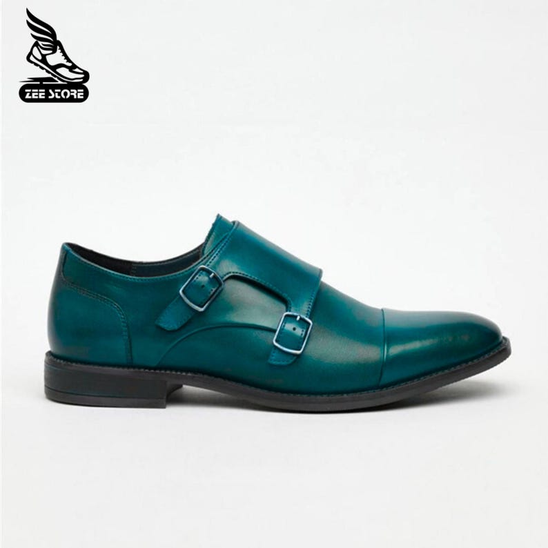 May include: Teal leather double monk strap dress shoe with a cap toe. The shoe features two adjustable buckles and a black sole. The Zee Store logo is in the upper left corner.