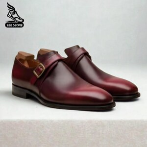 May include: A pair of burgundy leather dress shoes with a single strap and buckle closure. The shoes have a polished finish and a classic design, suitable for formal occasions. The Zee Store logo is in the upper left corner.