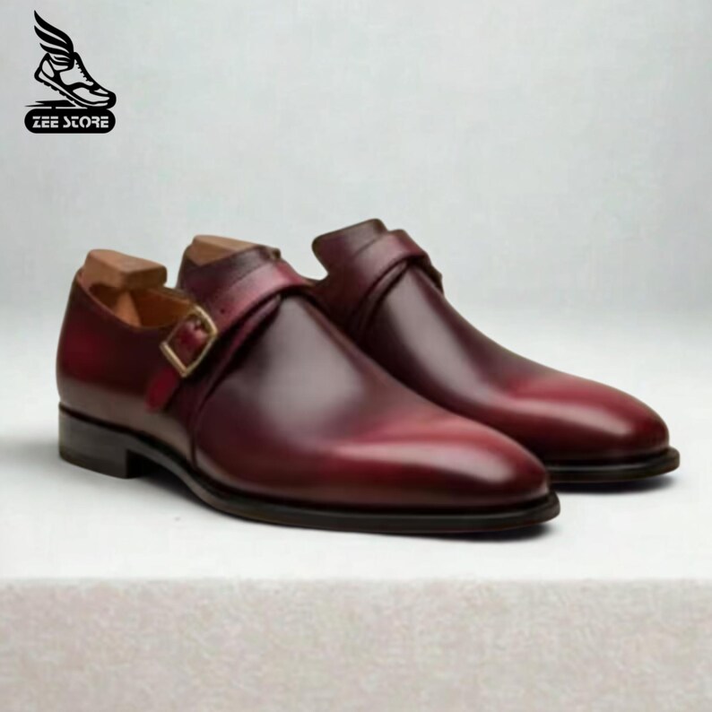 May include: A pair of burgundy leather dress shoes with a single strap and buckle closure. The shoes have a polished finish and a classic design, suitable for formal occasions. The Zee Store logo is in the upper left corner.