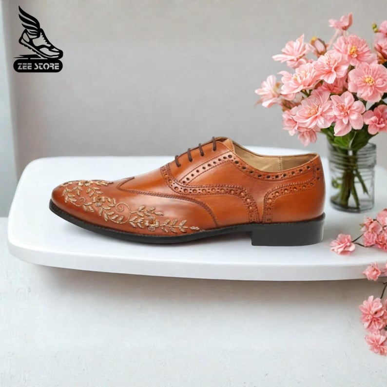 May include: A brown leather oxford shoe with decorative floral embroidery on the toe. The shoe features a wingtip design with brogue detailing and brown laces. The shoe is displayed on a white surface with a vase of pink flowers.