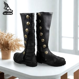 Puede incluir: Un par de botas negras hasta la rodilla con botones decorativos dorados y cordones. Las botas tienen una punta redonda y una suela resistente. Las botas se exhiben sobre una superficie blanca.