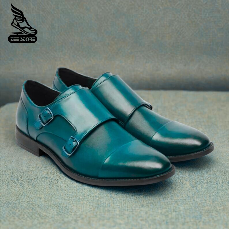 May include: A pair of teal leather dress shoes with double monk straps and silver buckles. The shoes have a glossy finish and a classic design, perfect for formal occasions. The soles are black.