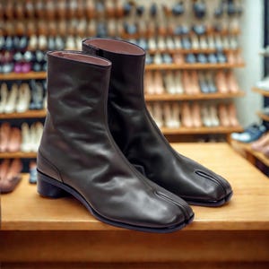 May include: A pair of dark brown leather ankle boots with a split-toe design. The boots have a smooth texture, a low heel, and a sleek, modern silhouette. The boots are displayed on a wooden surface, with a blurred background of shoe shelves.