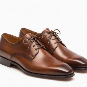 Op de afbeelding: Een paar bruine leren herenschoenen met een klassiek ontwerp. De schoenen hebben een gepolijste afwerking, veters en een lage hak. Het leer is een rijke, warme bruine kleur en de schoenen staan tegen een witte achtergrond.