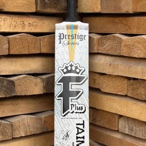 May include: A cricket bat with a black handle and a white and black grip. The bat face features the text "Prestige Series", a crown logo, and the text "F Plus". The name "TAIMOOR" is printed vertically on the bat.