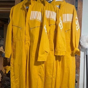 May include: Three yellow coveralls hanging on a rack. Each coverall has a barcode design and the numbers "42617 00002" printed on the chest. The coveralls have long sleeves and zippers.
