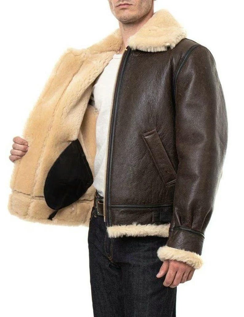 K&ouml;nnte beinhalten: Eine dunkelbraune Leder-Bomberjacke mit dickem, cremefarbenem Lammfellfutter und Kragen. Die Jacke verf&uuml;gt &uuml;ber einen Frontrei&szlig;verschluss aus Metall, lange &Auml;rmel mit Lammfellb&uuml;ndchen und zwei Seitentaschen. Sie wird &uuml;ber einem wei&szlig;en Hemd getragen.