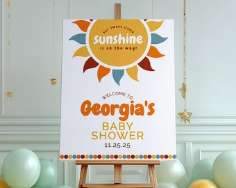 May include: A baby shower sign on a wooden easel. The sign has a sun design with the text "our sweet little sunshine is on the way!" and "Welcome to Georgia's BABY SHOWER 11.25.25" in orange and brown.