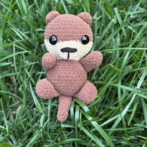 May include: A handmade crochet otter plush toy in shades of brown, cream, and black, with a small tail and outstretched arms. The plush is set against a backdrop of green grass.