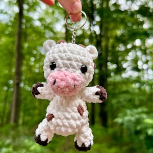 Puede incluir: Un llavero de vaca de ganchillo blanco hecho a mano con manchas marrones, pezuñas y detalles en las orejas. La vaca tiene un hocico rosa y ojos negros. El llavero se sujeta con un anillo y una cadena plateados.