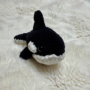 May include: A handmade plush orca whale in black and white. The orca has a black body with white patches around the eyes and belly. The toy is lying on a white, fluffy surface.