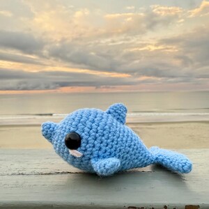 May include: A light blue crocheted dolphin toy with black eyes and a small white detail. The handmade plushie is lying on a wooden surface, with a beach and sunset in the background.
