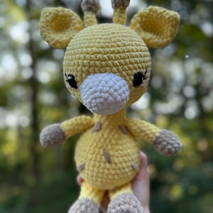 Puede incluir: Un juguete de jirafa de ganchillo hecho a mano en tonos amarillo, blanco y marrón. La jirafa tiene orejas grandes y redondas, pequeños cuernos y un hocico blanco. El juguete es suave y afelpado, con costuras detalladas y una expresión amigable.