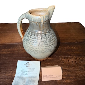 May include: A stoneware pitcher with a rounded body, a curved handle, and a textured, mottled blue-grey glaze. The pitcher is accompanied by a white informational card from Browning Artworks and a small tan card labeled Brad Tucker Functional Stoneware Pottery.