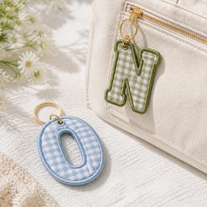May include: A cream-colored canvas tote bag with a gold zipper, featuring a green and white gingham letter N keychain attached to the zipper pull, and a separate blue and white gingham letter O keychain resting nearby on a textured surface.