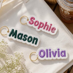 May include: Three embroidered fabric keychains with gold-tone metal rings. Each keychain features a white scalloped border and a name stitched in a different colour: pink for Sophie, dark green for Mason, and purple for the name Olivia.
