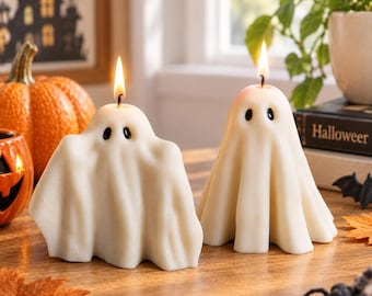 May include: Two off-white ghost-shaped candles with black painted eyes and lit wicks. The candles have a draped, fabric-like texture and are positioned on a wooden surface, creating a festive display with a warm, flickering flame on each.