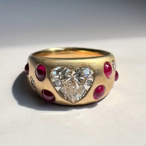 May include: A gold ring featuring a heart-shaped diamond center stone. The ring is adorned with round, red gemstones and has a brushed finish. The ring is a piece of jewelry.