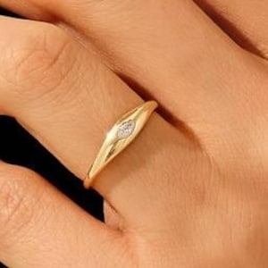 May include: A gold ring with a small, oval-shaped diamond set in the center. The ring is a simple, elegant design, with a smooth, polished finish. The ring is on a finger.