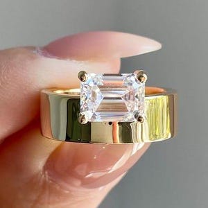 Op de afbeelding: Een gouden ring met een grote, rechthoekige, heldere edelsteen. De ring heeft een brede, platte band. De edelsteen is gezet in een vierkante zetting. De ring wordt door een hand omhoog gehouden.