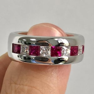 Peut inclure: Une bague en argent avec un sertissage en canal, ornée d'alternances de pierres rouges carrées et de pierres transparentes. La bague est tenue par un doigt, mettant en valeur le métal poli et les couleurs vives des pierres.