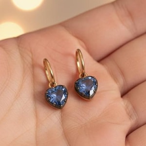 Könnte beinhalten: Ein Paar goldene Creolen, jeweils mit einem herzförmigen blauen Edelstein. Die Ohrringe werden in einer offenen Hand gehalten, vor einem weichen, verschwommenen Hintergrund aus warmen, unscharfen Lichtern.