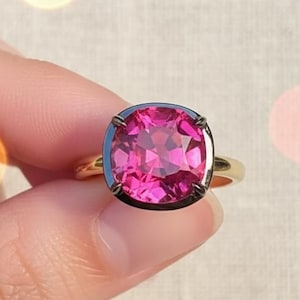 Könnte beinhalten: Ein goldener Ring mit einem großen, kissenförmig geschliffenen, leuchtend rosa Edelstein. Der Edelstein ist in einer dunklen Lünette gefasst, was einen Kontrast erzeugt. Der Ring hat ein poliertes Goldband.