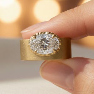 Op de afbeelding: Een gouden ring met een brede band en een ovaalvormig middelpunt. Het middelpunt heeft een grote, heldere edelsteen omringd door kleinere stenen. De ring wordt tussen vingers gehouden tegen een vage achtergrond.