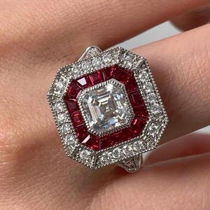 Peut inclure: Bague carrée en diamant avec un halo de rubis et de diamants. La bague présente un diamant central de taille carrée entouré d'un halo de petits diamants et d'un second halo de rubis rouges. La bague est sertie dans un métal argenté.