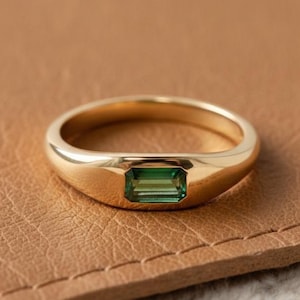 May include: A gold ring with a rectangular, emerald-green gemstone set in a bezel. The ring has a smooth, polished band and a classic, minimalist design. The ring is displayed on a brown leather surface.