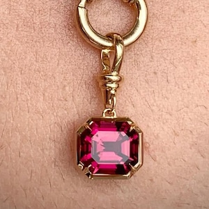 May include: A gold-colored necklace with a square, ruby-colored pendant. The pendant is set in a gold frame and hangs from a gold clasp. The necklace chain is made of small, round beads.