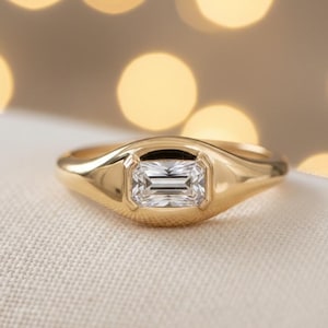 May include: A gold ring with a rectangular, clear gemstone set in the center. The ring has a smooth, polished band and a bezel setting that holds the gemstone securely. The background is blurred with soft, golden bokeh.