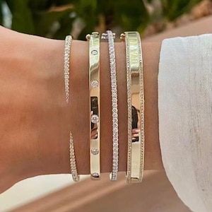 Puede incluir: Una colección de pulseras de oro y diamantes. Las pulseras incluyen una fina pulsera de tenis de diamantes, una pulsera de oro con diamantes y una pulsera de oro más ancha con un borde de diamantes. Las pulseras se llevan en una muñeca.