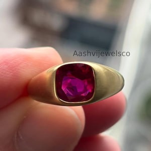May include: A gold-toned ring featuring a large, square-cut, deep red gemstone. The ring has a smooth, polished band and a bezel setting that securely holds the vibrant stone. The ring is held in a hand, with the brand name "Aashvijewelsco" visible.