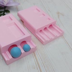 May include: Two pale pink moulds with different shaped cavities. One mould has two blue spheres inside. The other mould has three curved grooves. The moulds are on a light wood surface with a purple flower in the background.