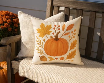 May include: A decorative fall-themed pillow with a pumpkin and autumn leaves design. The pillow is cream-colored with an orange pumpkin, yellow leaves, and brown accents. It rests on a wooden bench with a cream-colored blanket.