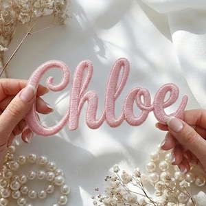 以下が含まれることがあります： ピンク色の刺繍パッチで、筆記体で「Chloe」と書かれています。白い布の背景に、2つの手でパッチが持たれており、手前には真珠とドライフラワーがあります。