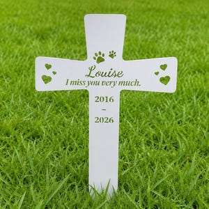 May include: A white cross-shaped memorial marker featuring the name Louise, the text I miss you very much, the years 2016-2026, two paw prints, and four heart-shaped cutouts. The marker is designed to be inserted into the ground.