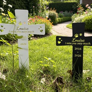 May include: Two metal cross-shaped garden markers, one white and one black, featuring cut-out heart and paw print designs. Each cross is engraved with the name Louise, the phrase I miss you very much, and the years 2016 - 2026 in gold lettering.