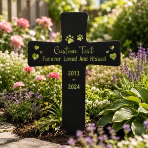 May include: A black metal cross garden stake featuring laser-cut details including two paw prints at the top, four heart-shaped cutouts, and engraved text reading Custom Text, Forever Loved And Missed, 2011, and 2024 in a gold-toned finish.