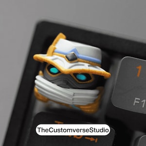 Op de afbeelding: Een aangepaste keycap met een karakterontwerp met een witte hoed, blauwe accenten en gouden rand. De keycap heeft gloeiende blauwe ogen en een zwart gezichtsmasker. De tekst "TheCustomverseStudio" is zichtbaar op het toetsenbord.