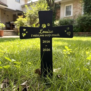 May include: A black cross-shaped memorial marker featuring gold-colored text that reads Louise, I miss you very much, and the years 2016-2026. The design includes cut-out heart shapes and paw prints at the top, with a stake for ground insertion.