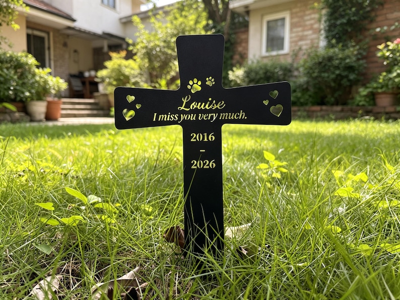 May include: A black cross-shaped memorial marker featuring gold-colored text that reads Louise, I miss you very much, and the years 2016-2026. The design includes cut-out heart shapes and paw prints at the top, with a stake for ground insertion.