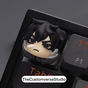 Può includere: Un copritasto nero personalizzato raffigurante un personaggio in stile anime con capelli neri e una benda. Il copritasto è posizionato su una tastiera nera, con il testo "TheCustomverseStudio" visibile. Un accessorio unico.