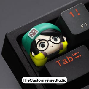 May include: A custom keycap featuring a cartoon character with a green hat, glasses, and yellow accents. The keycap is set on a black keyboard, with the "Tab" and "1!" keys visible. TheCustomverseStudio is printed on the bottom.