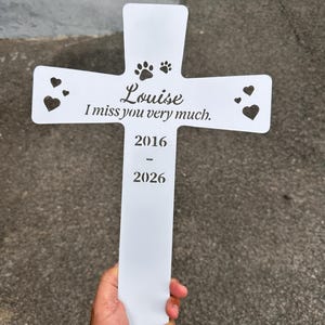 May include: A white metal cross-shaped garden stake featuring cut-out heart and paw print designs. The text reads Louise, I miss you very much, 2016, and 2026. The surface is smooth and the design is centered on the vertical and horizontal bars.